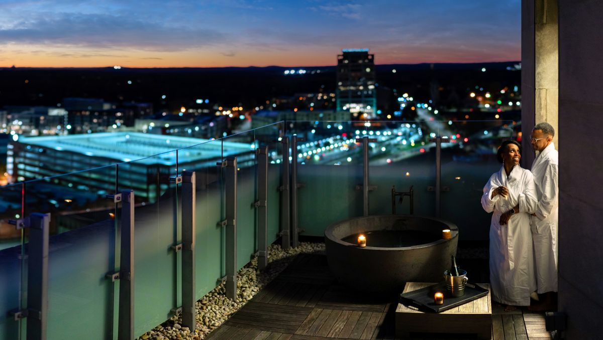 Man and woman standing in white robes beside private soaking tub on terrace overlooking Durham at night