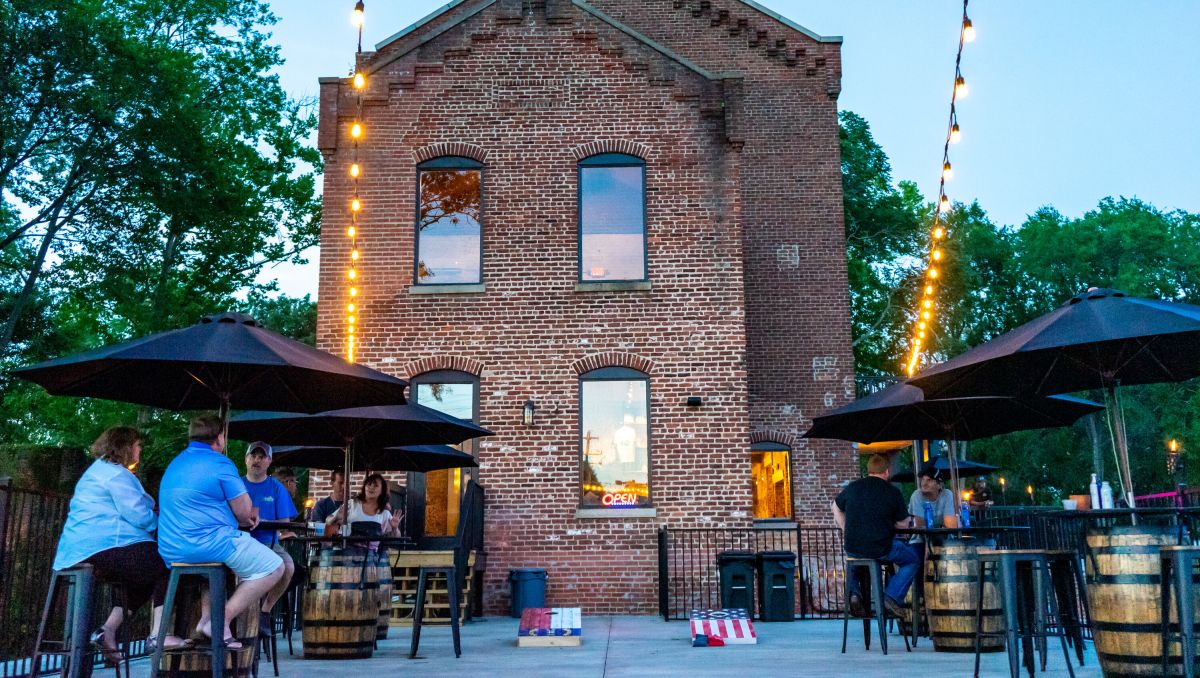 People enjoying spirits under umbrellas on Weldon Mills Distillery patio with two-story brick building in background