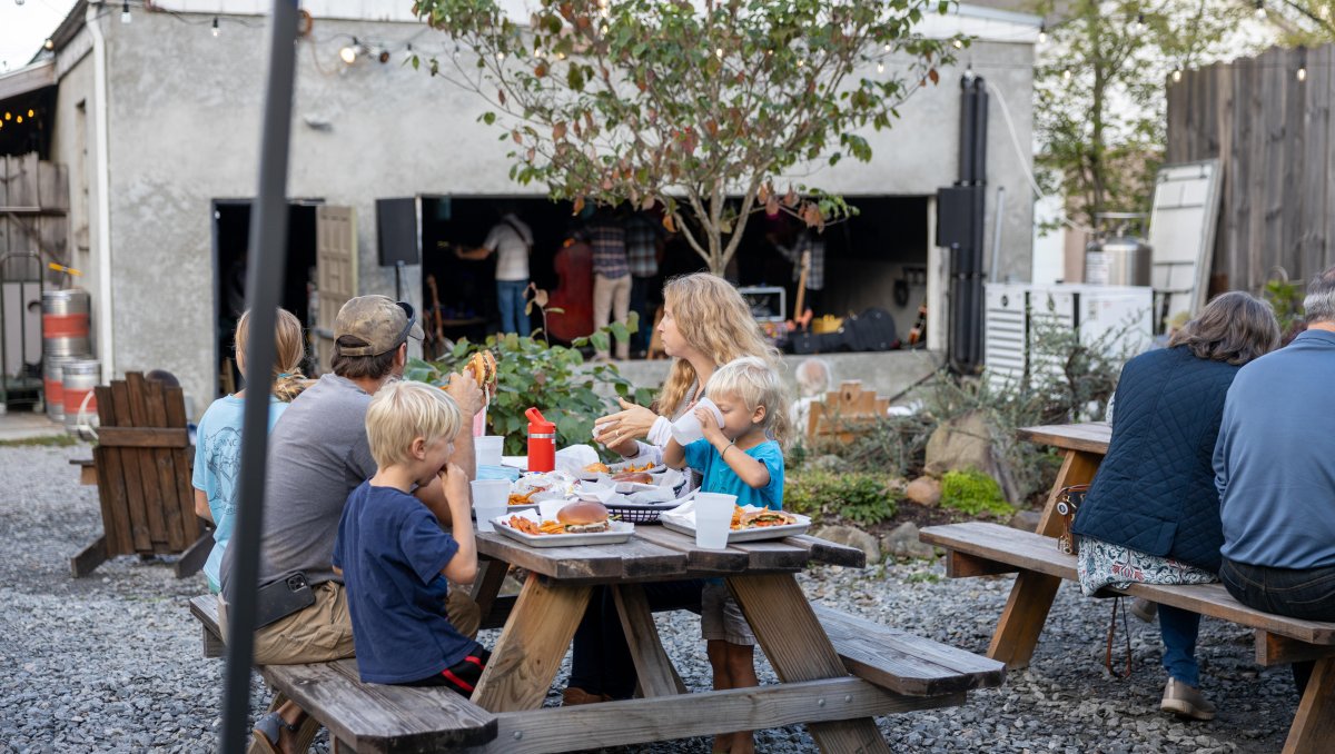 People sitting at picnic tables eating food with band playing in background inside building