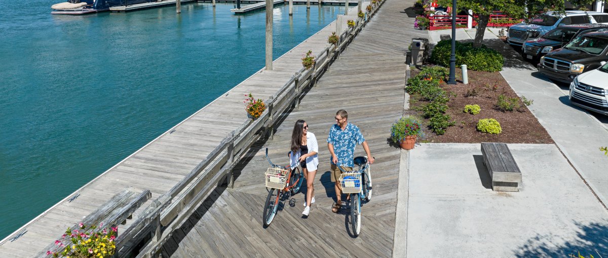 Couple walking bikes down wood Riverwalk with quaint waterfront and boats in background