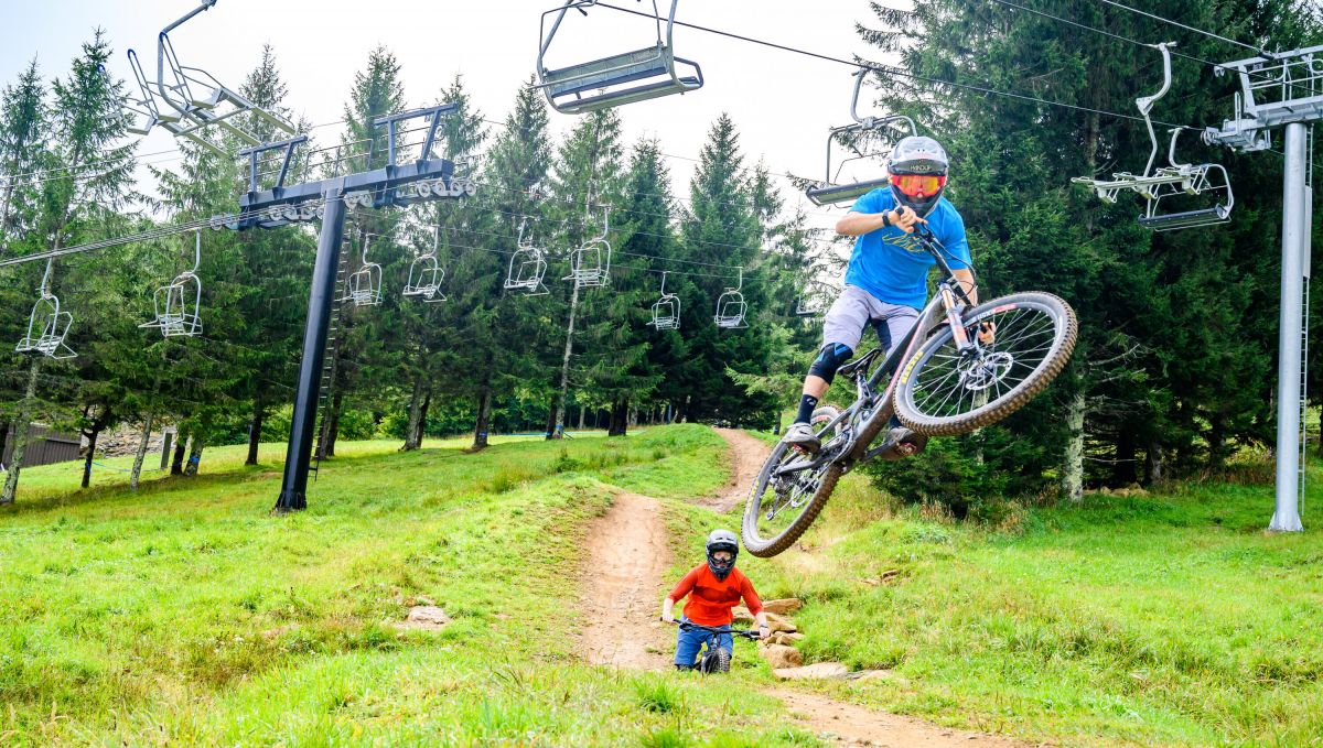 Two friends mountain biking in Beech Mountain, and the one in front getting air, with ski lifts above them