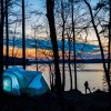 Silhouette of person fishing in lake at dusk by tent with sun setting in distance