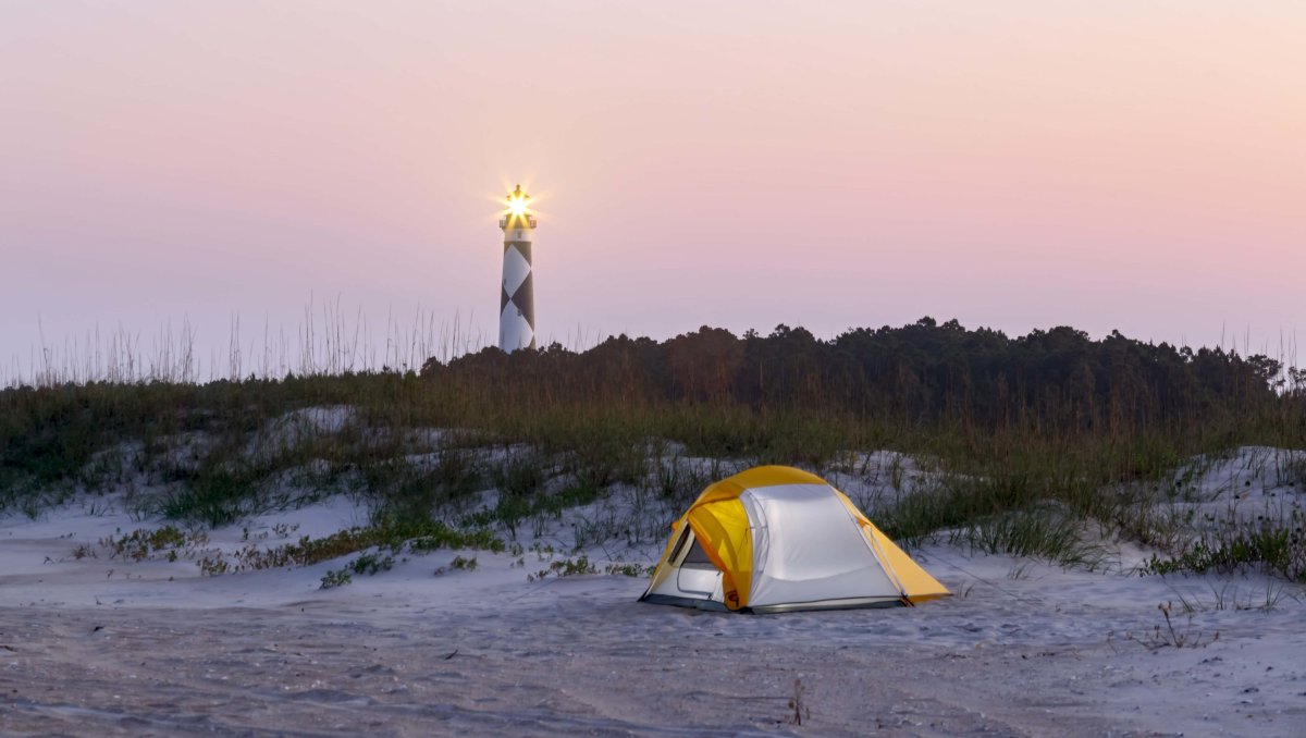 Small tent pitched on sand with dunes and lighthouse in background at dusk