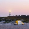 Small tent pitched on sand with dunes and lighthouse in background at dusk