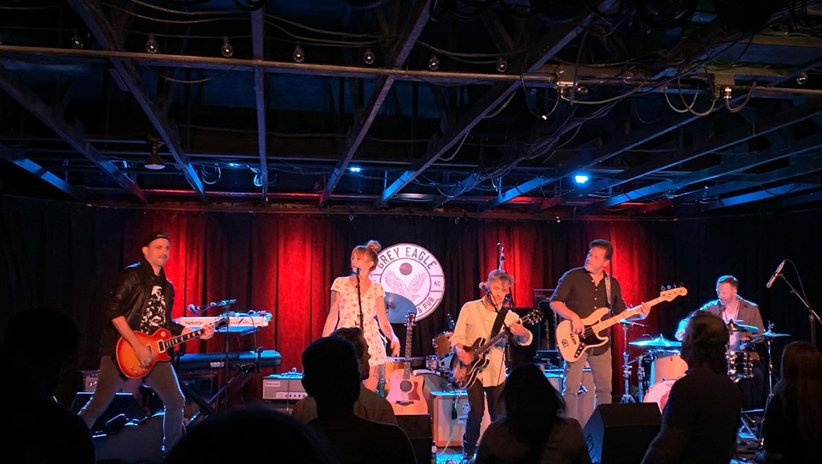 Band performing on stage with red lights in background in front of crowd indoors