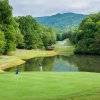 Golfer hitting shot near water onto green with pond, trees and mountains in background