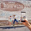 Phone in foreground taking photo of friends jumping in front of Cheerwine mural on side of building