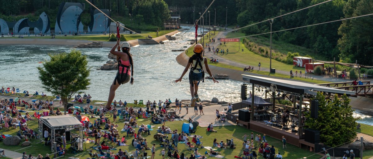 Two people going down zip lines with crowd of people on lawn below and whitewater river in background