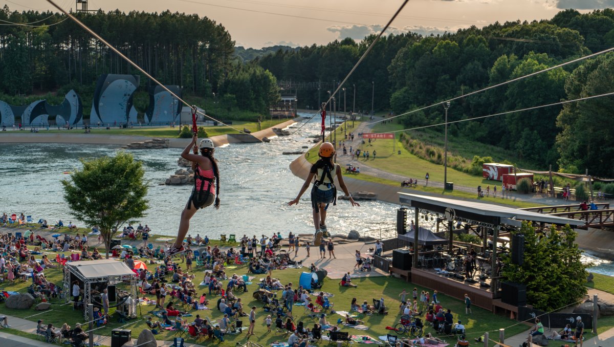 Two people going down zip lines with crowd of people on lawn below and whitewater river in background