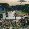 Two people going down zip lines with crowd of people on lawn below and whitewater river in background