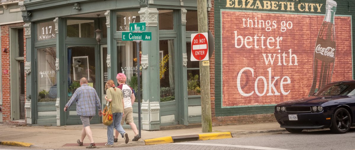 People walking through crosswalk in front of old buildings and a Coca-Cola mural