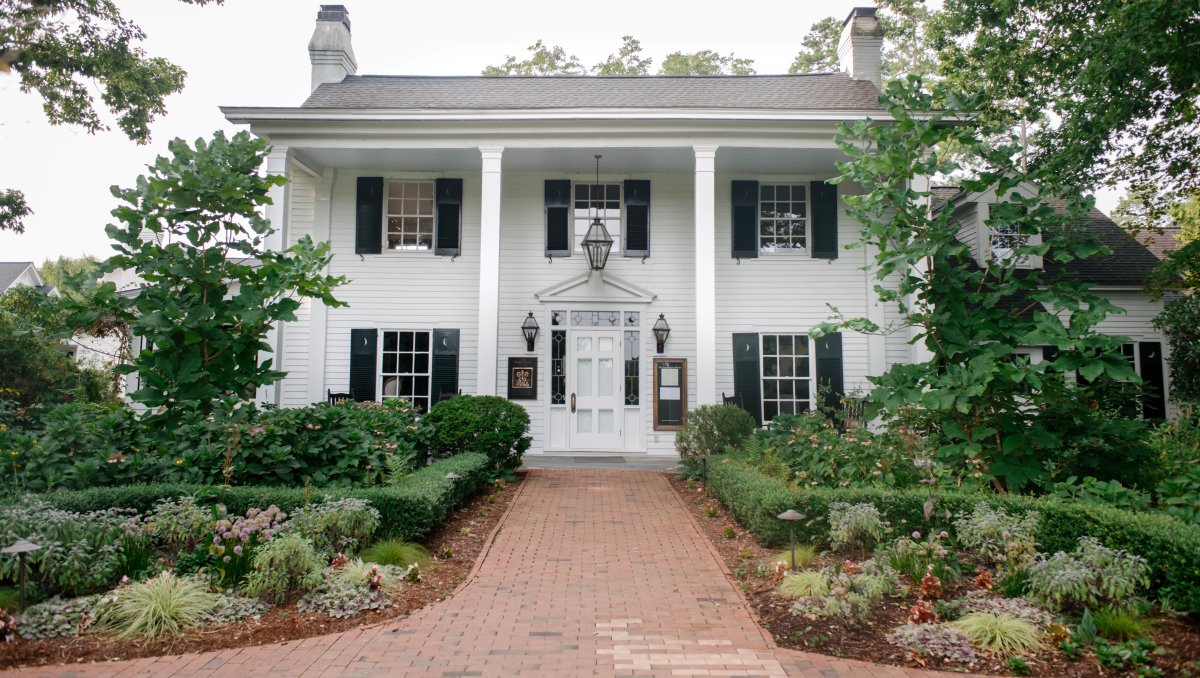 Front exterior of a beautiful white inn, with gardens and brick sidewalk in front during daytime