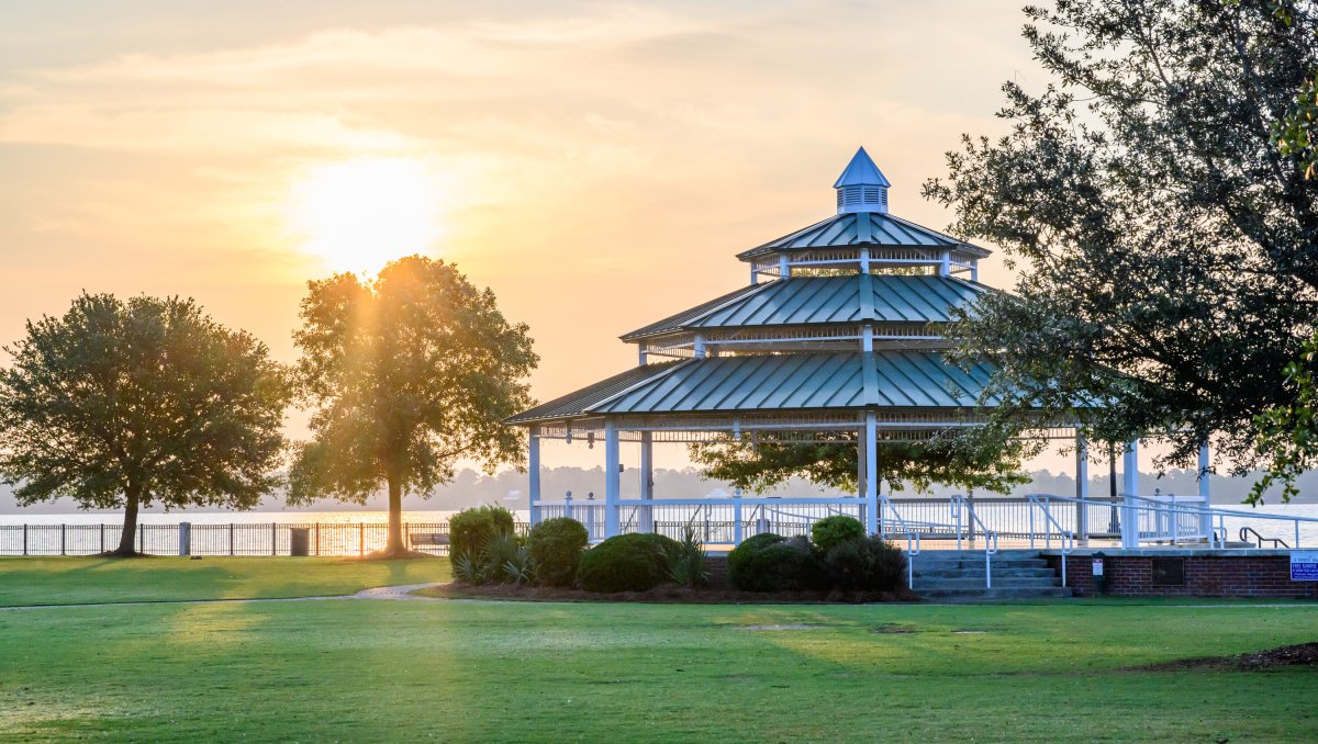 Large gazebo in middle of empty park surrounded by green grass and trees with sun shining