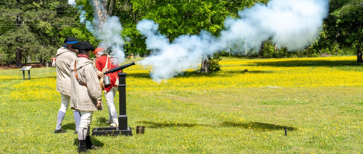 Interpreters shooting off cannon in field with trees in background