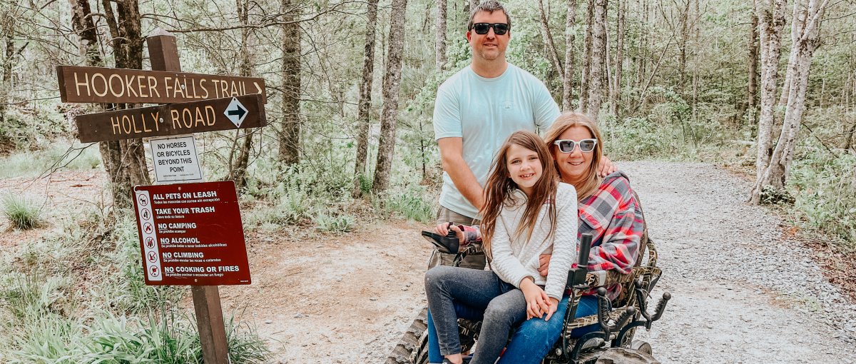 Woman in track outdoor chair posing with daughter and husband in front of waterfalls sign on trail in forest