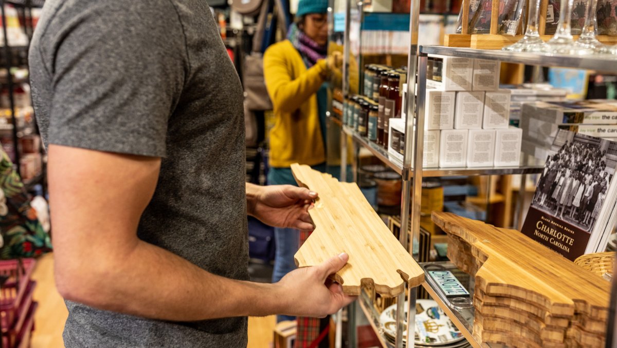 Man holding North Carolina-shaped cutting board in gift shop with woman looking at shelves of items in background