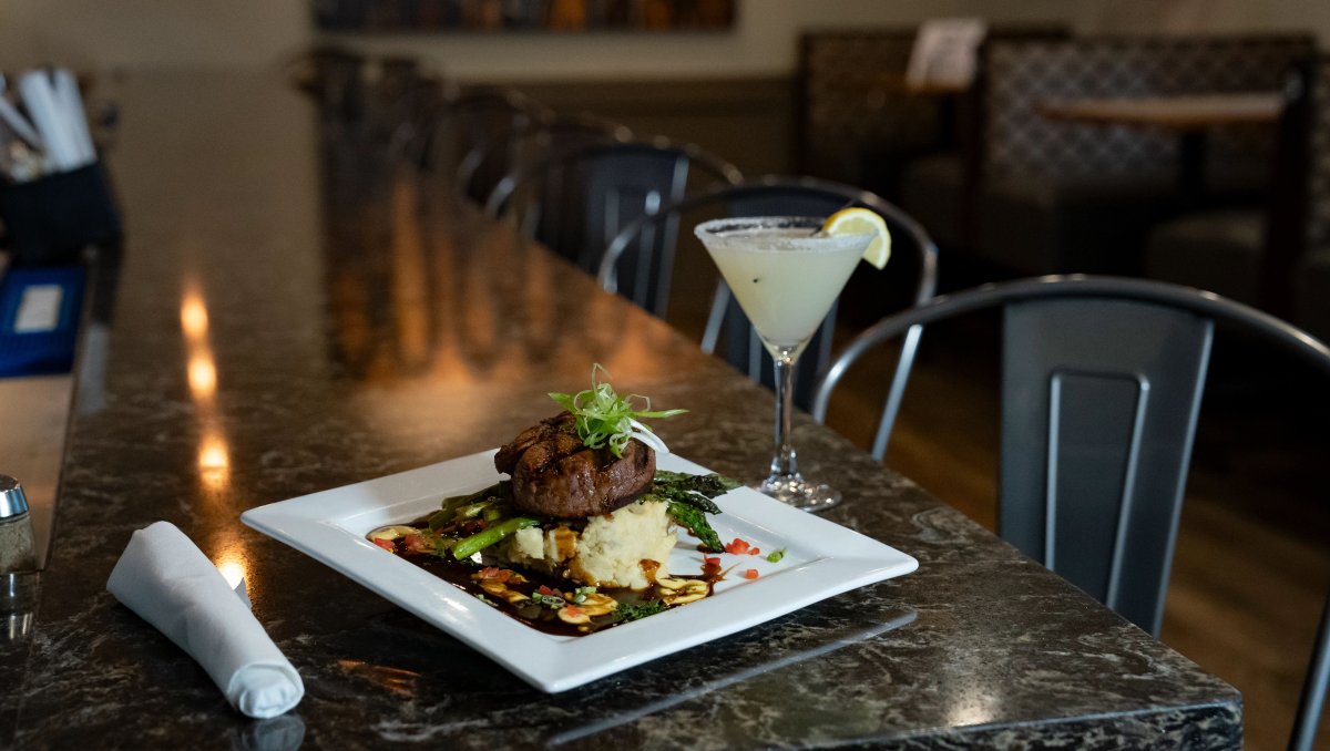 Plated steak dish sitting on restaurant's bar with lemon martini beside it