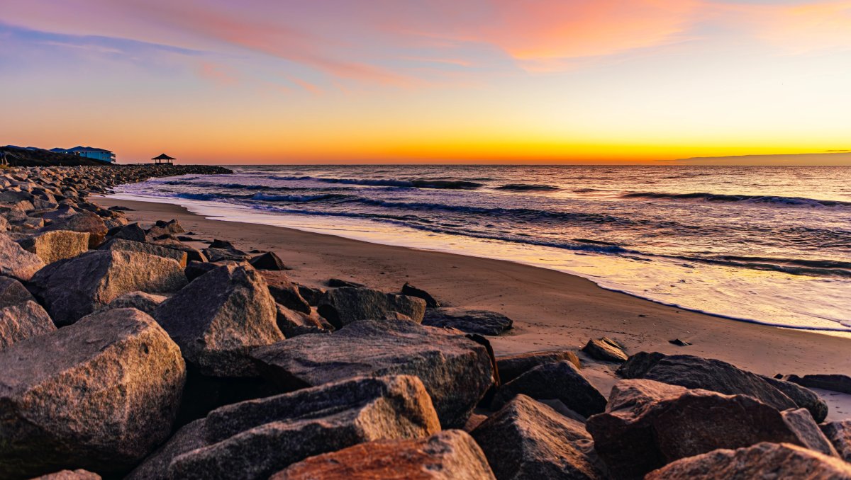 Sun rising over ocean on horizon with large rocks and sand in foreground