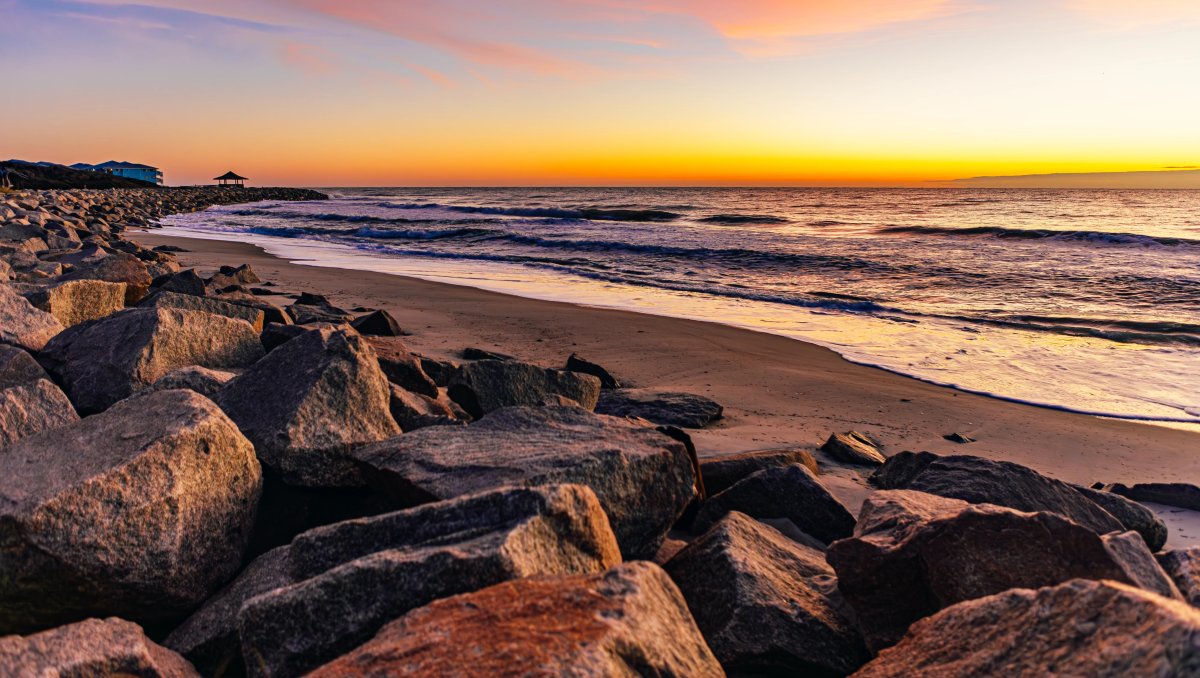 Sun rising over ocean on horizon with large rocks and sand in foreground