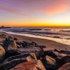 Sun rising over ocean on horizon with large rocks and sand in foreground