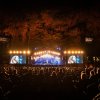 Performers on lit-up stage with trees in background and full crowd in foreground during nighttime