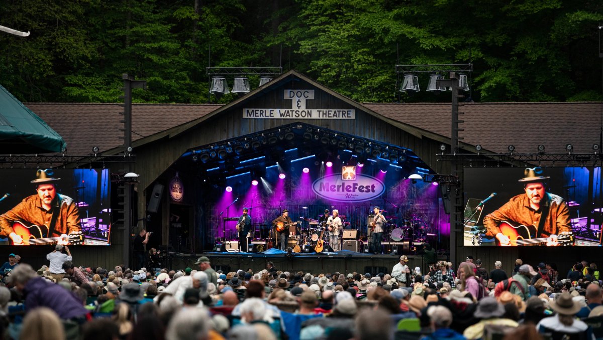 Band performing on stage under purple lights with large screens on both sides, trees in background and crowd in foreground