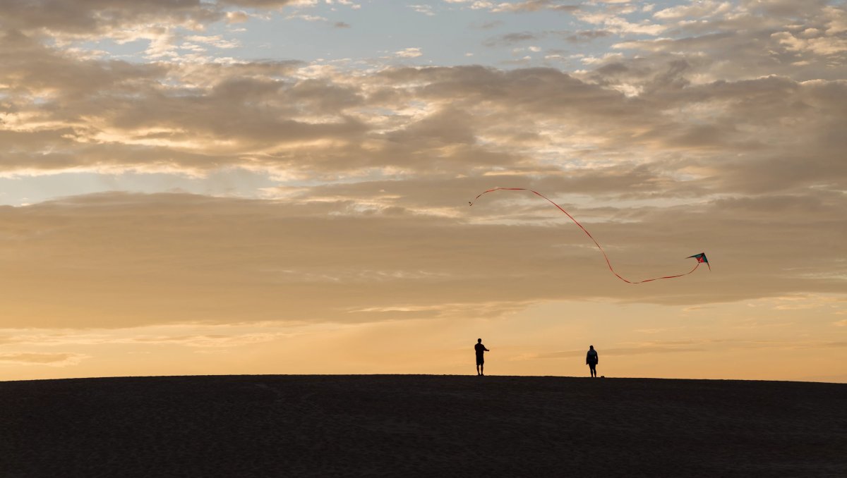 Silhouettes of two people flying a kite on sand dune, orange sky in background