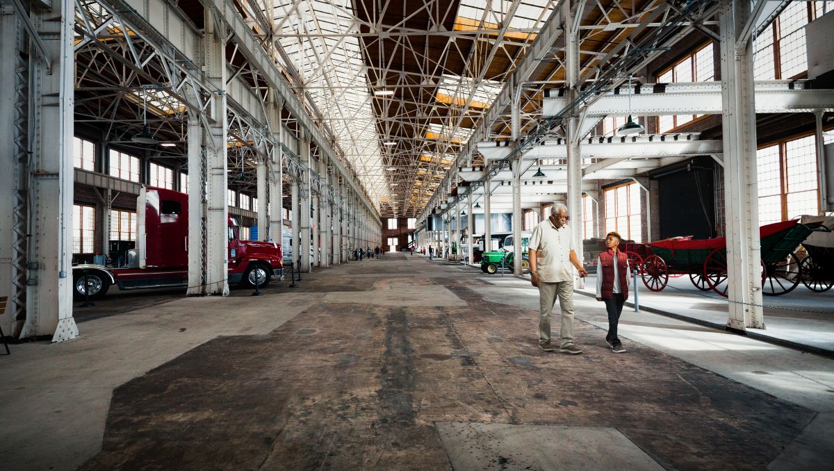 Man and child walking through NC Transportation Museum exhibits with automobiles and vast warehouse in background