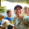 Smiling man taking a selfie with his wife, daughter and dog in front of a lake