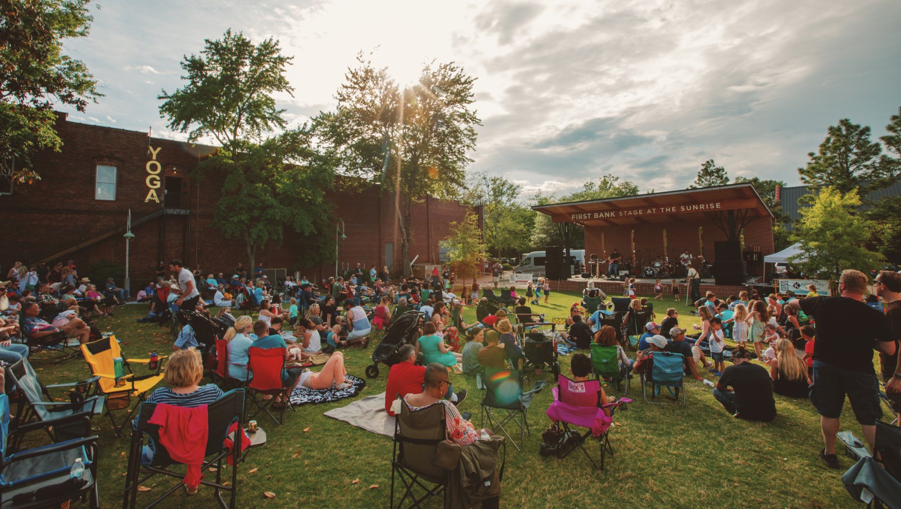 People in lawn chairs enjoying live music at small-town festival with sun setting