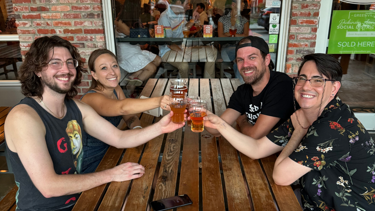 Four friends sitting at table cheersing beers and smiling at the camera