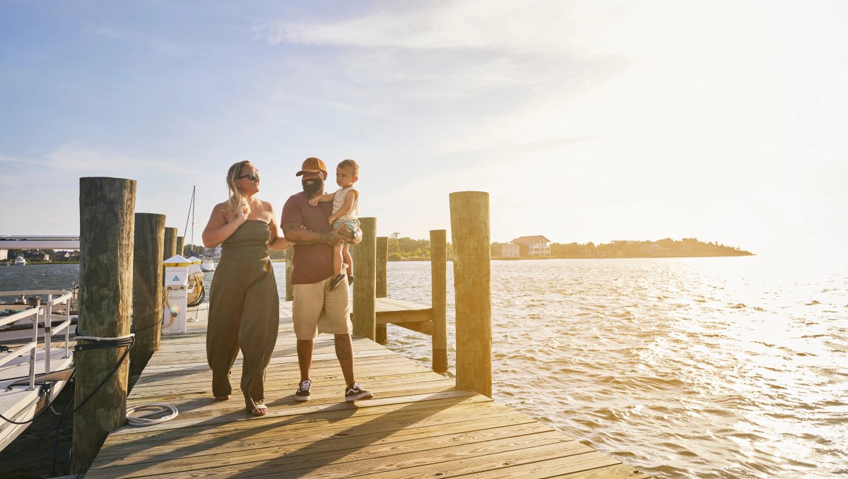 Family of 3 walking on dock over lake with sun and land in background