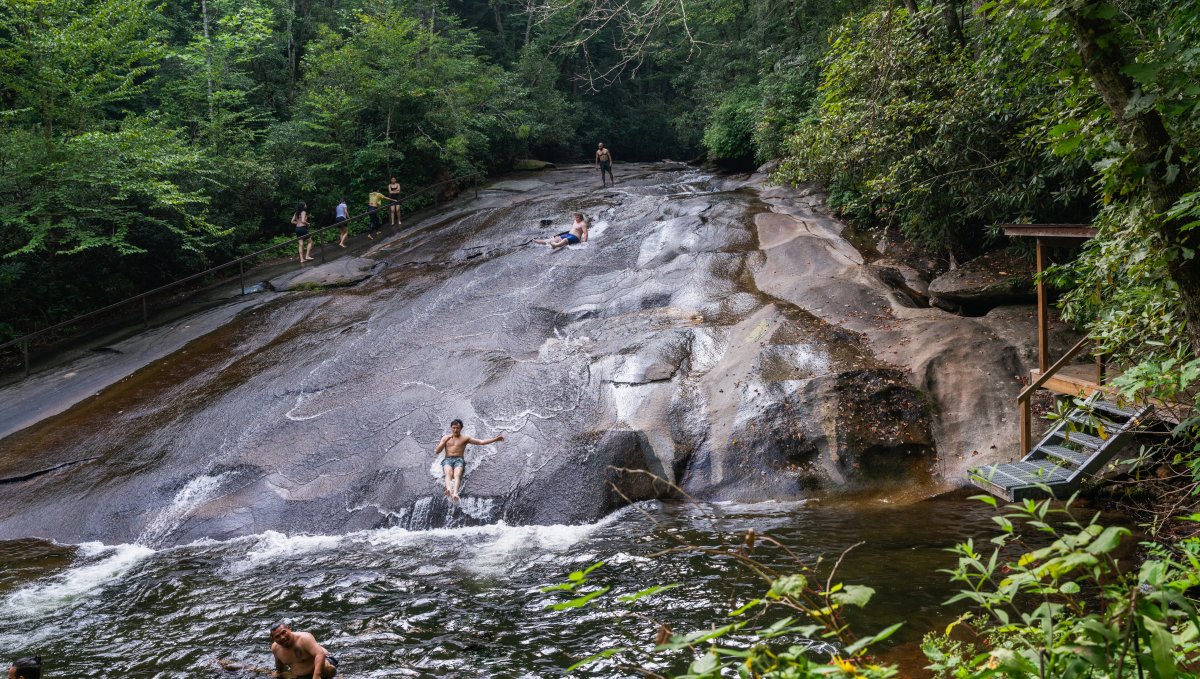 Kds sliding down rock into pool of natural water surrounded by green trees during daytime