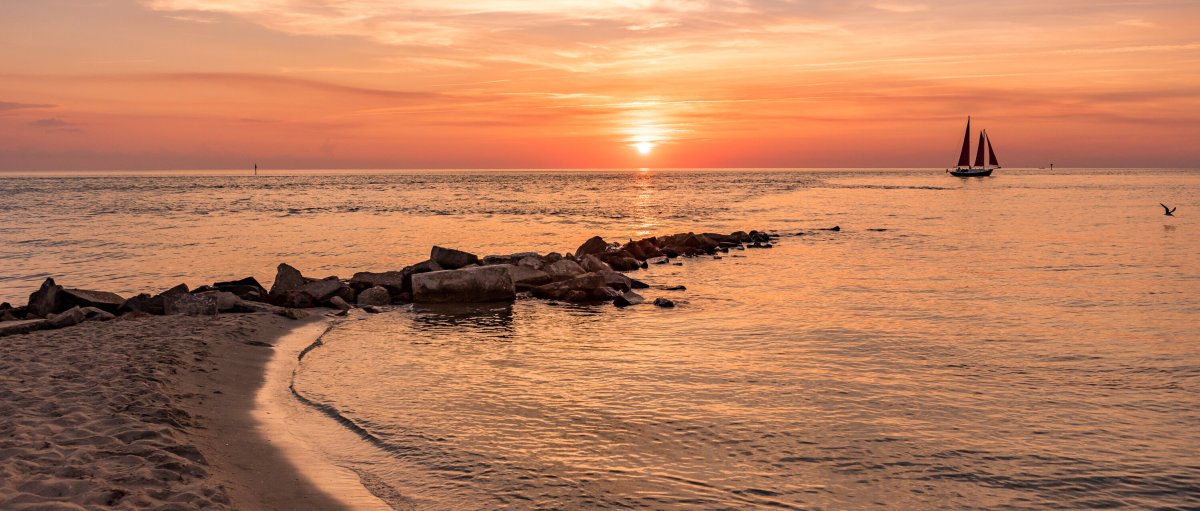 Rocks jutting out into ocean from beach with sailboat and sun setting in distance