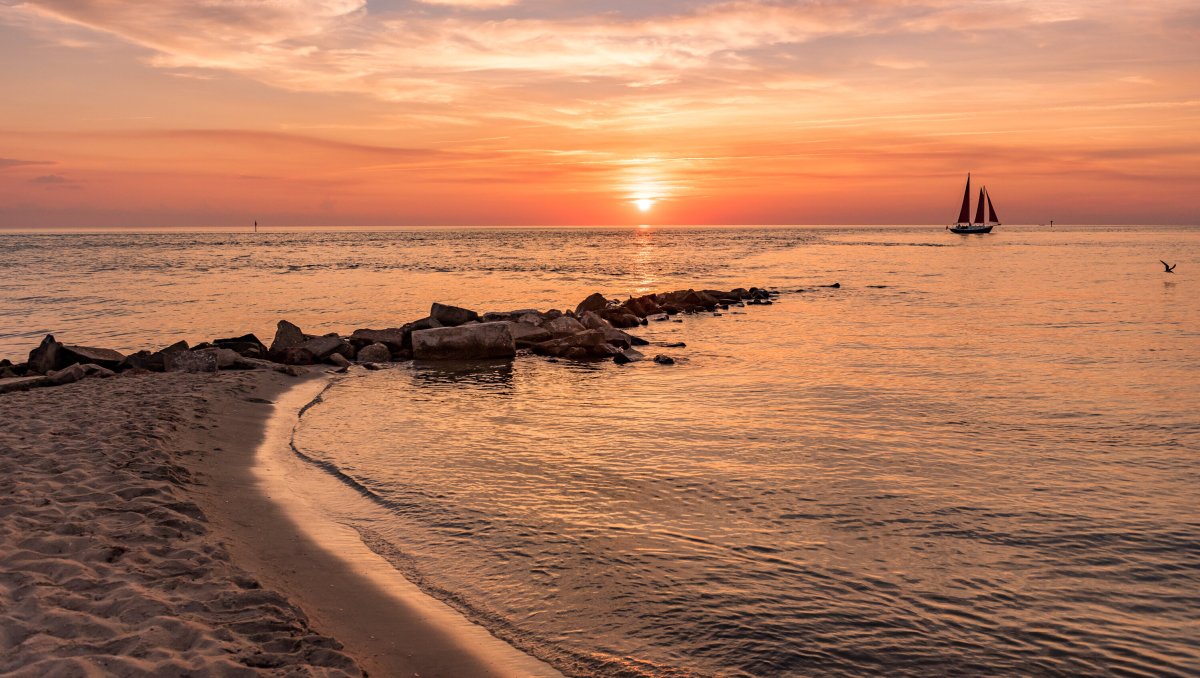 Rocks jutting out into ocean from beach with sailboat and sun setting in distance