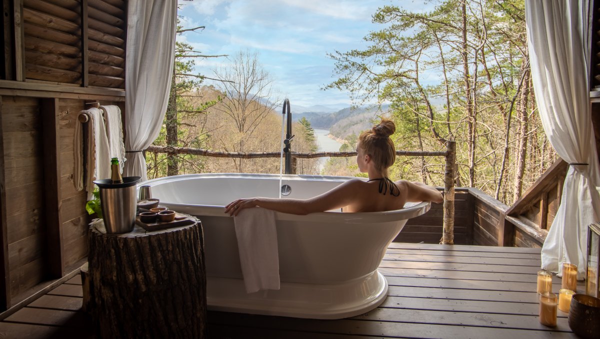 Woman in soaking tub in treehouse with forest, mountains and river in distance