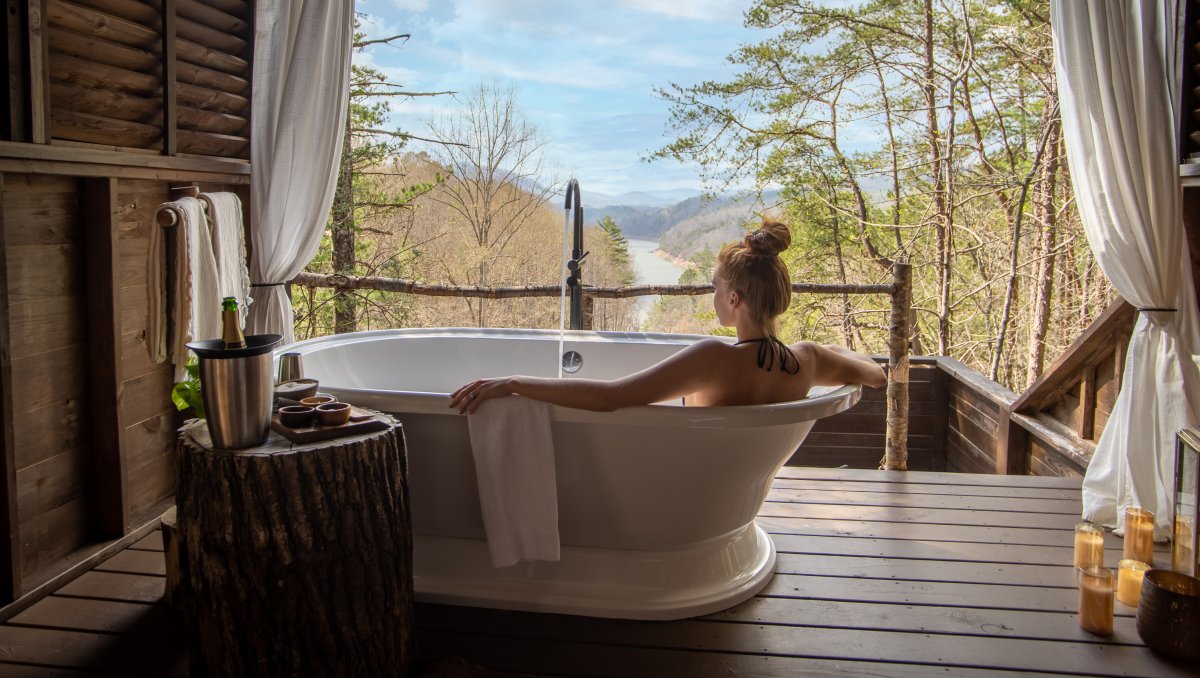 Woman in soaking tub in treehouse with forest, mountains and river in distance