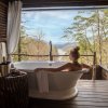 Woman in soaking tub in treehouse with forest, mountains and river in distance