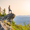 Woman standing on The Blowing Rock outcropping with green foliage below and mountains in background