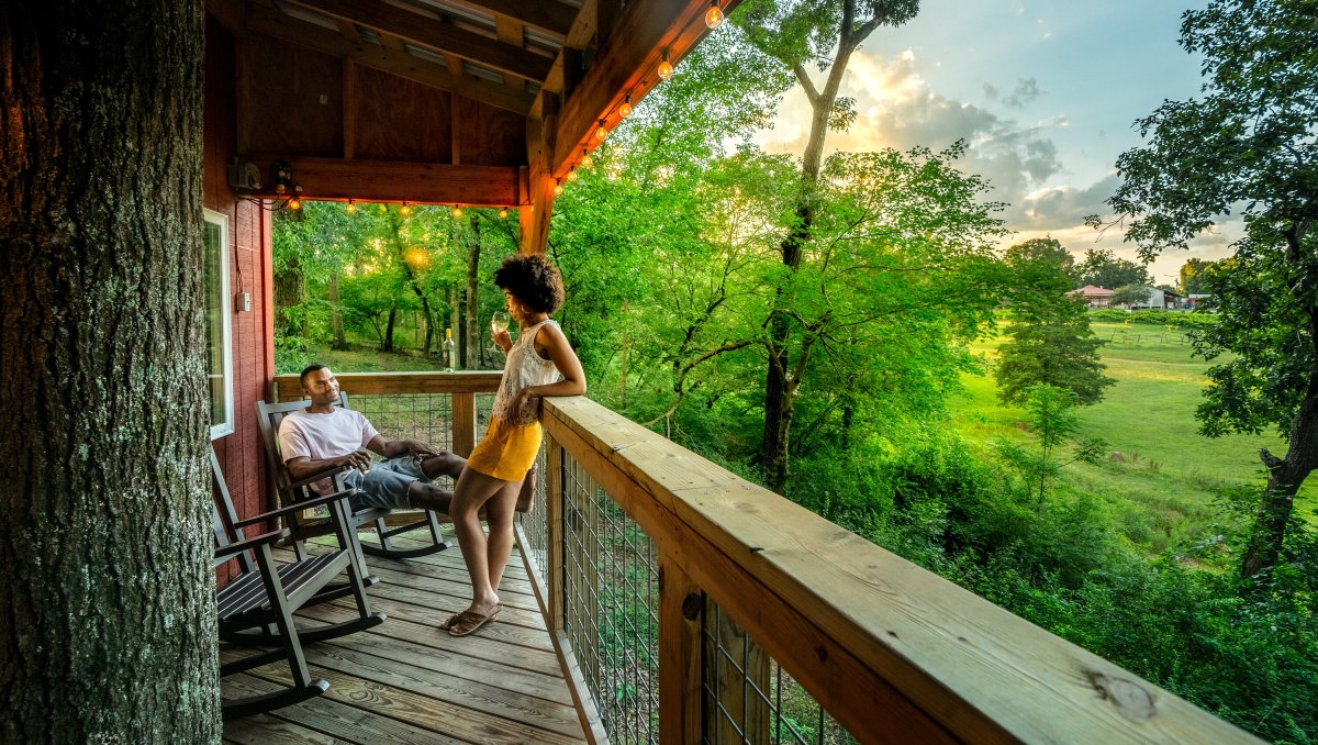 Couple enjoying wine on treehouse balcony with green trees and grounds to the right