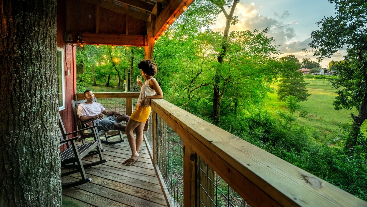 Couple enjoying wine on tree house balcony with green trees and grounds to the right