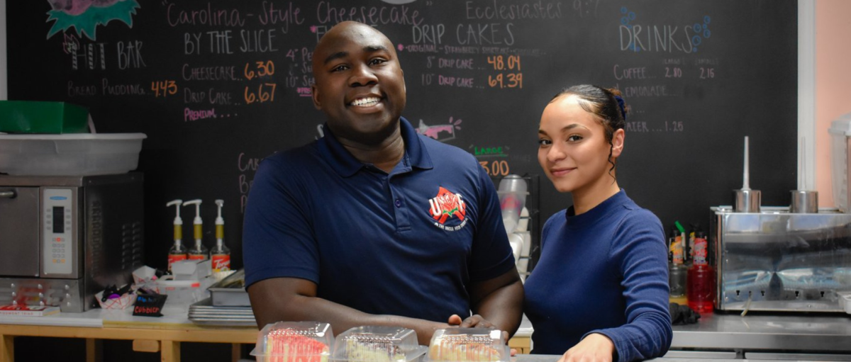 Two workers at cheesecake store smiling at camera with chalkboard behind them