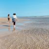 Two women walking on beach with calm, clear beach water at their feet with blue sky in background