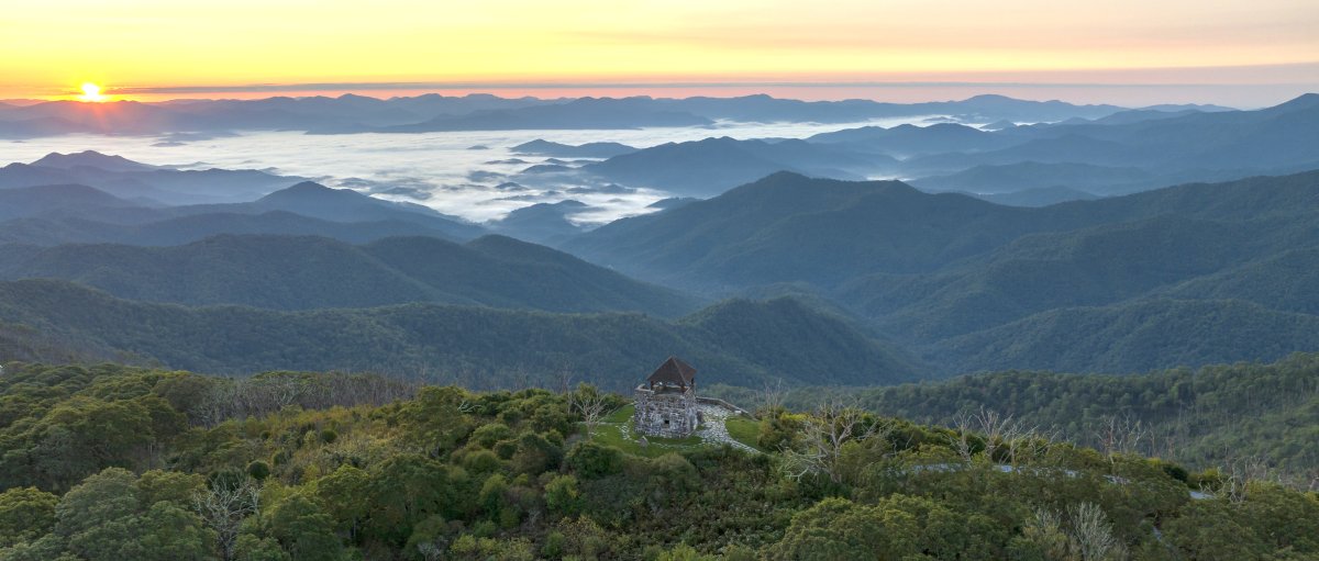 Aerial view of tower in middle of trees and mountains with sun rising in background