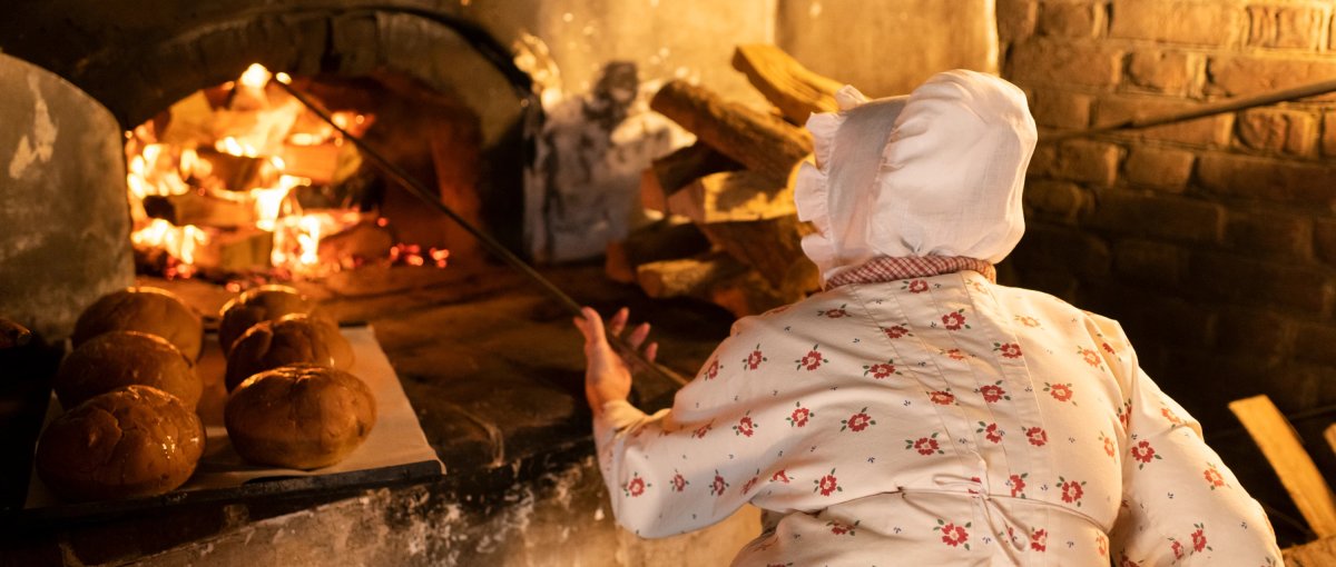 Back of Moravian baker inserting poker into wood fire at bakery