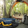 woman standing and boy sitting at picnic table in woods as seen from inside tent with camping supplies in foreground